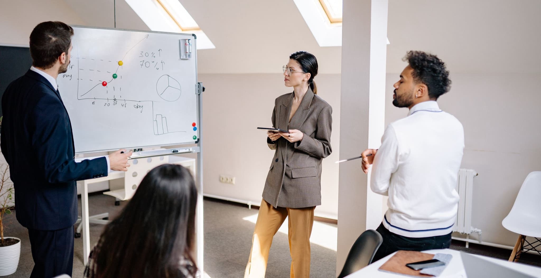 Consulting team in a bright office reviewing a hand-drawn roadmap chart with milestone markers on a whiteboard while a presenter holding a tablet leads the discussion