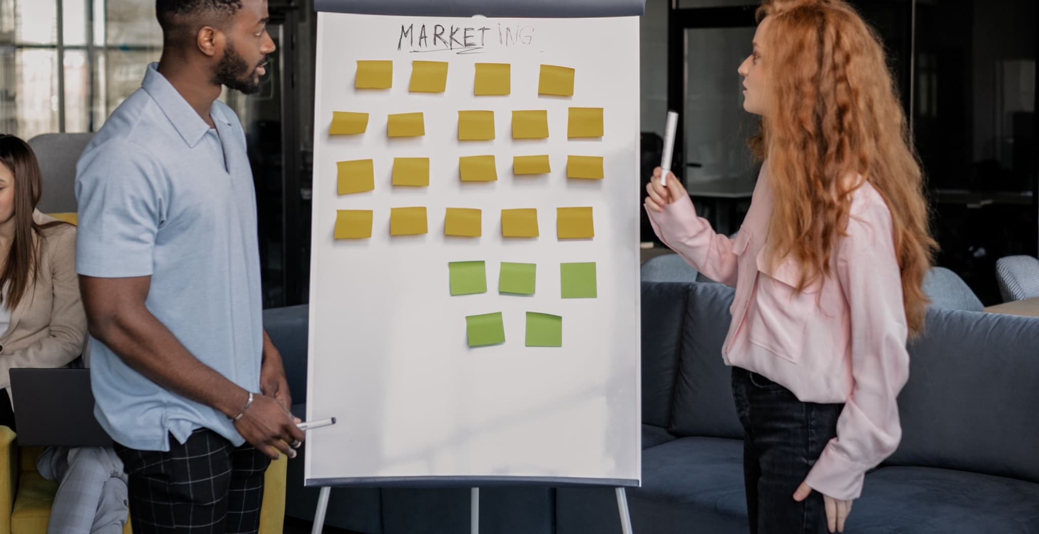 Two colleagues standing either side of a flipchart whiteboard covered in yellow and green sticky notes during a discovery workshop in a modern lounge office