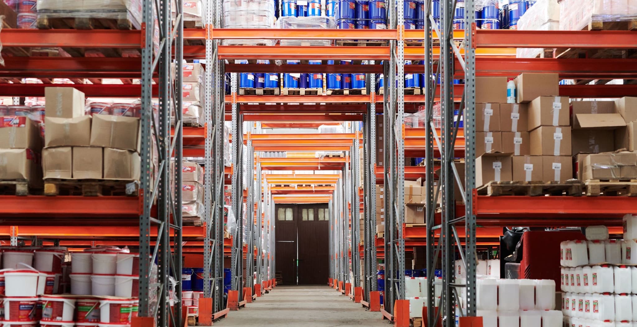 Symmetrical view down a wide warehouse aisle between tall pallet-racking bays stocked with shrink-wrapped pallets and boxes, leading to a dark double door at the far end