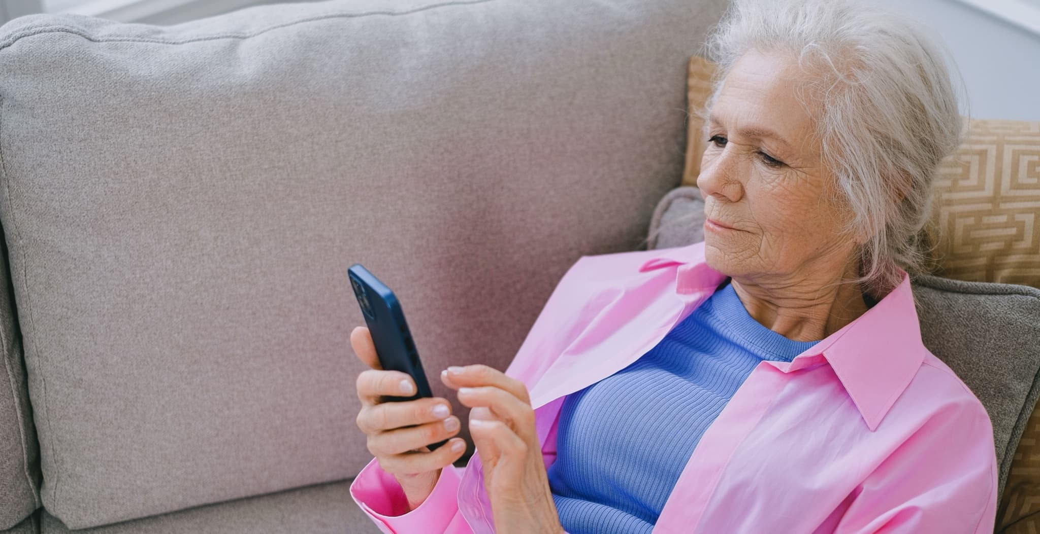 Older woman relaxing on a grey sofa at home, holding and browsing on her smartphone in soft daylight
