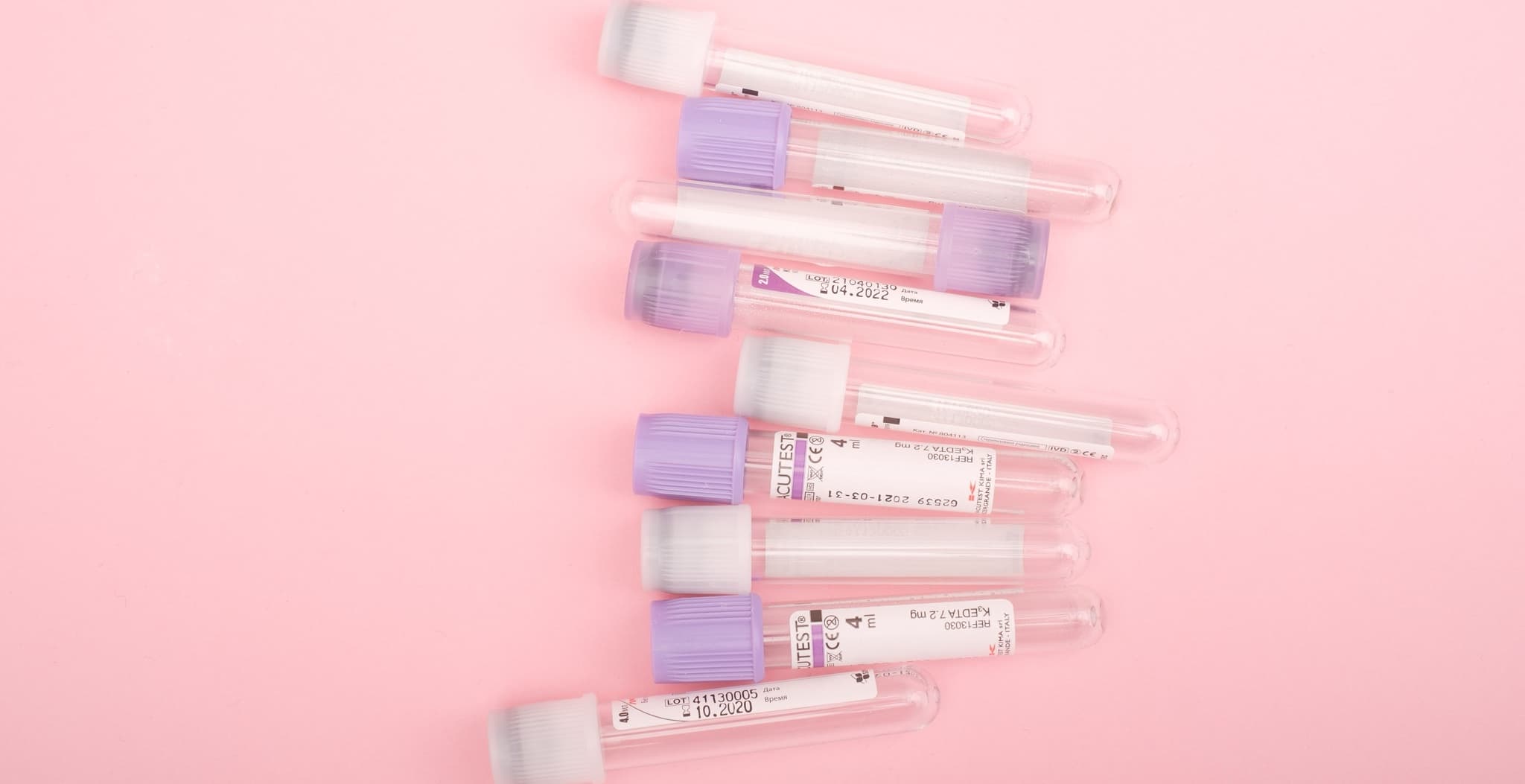 Rows of blood collection tubes with purple and white caps, several printed with 'ACUTEST' labels, arranged diagonally on a pink background.