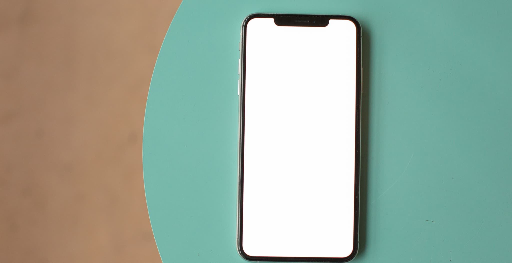 A smartphone with a blank white screen lying face-up on a table split between tan and teal surfaces.