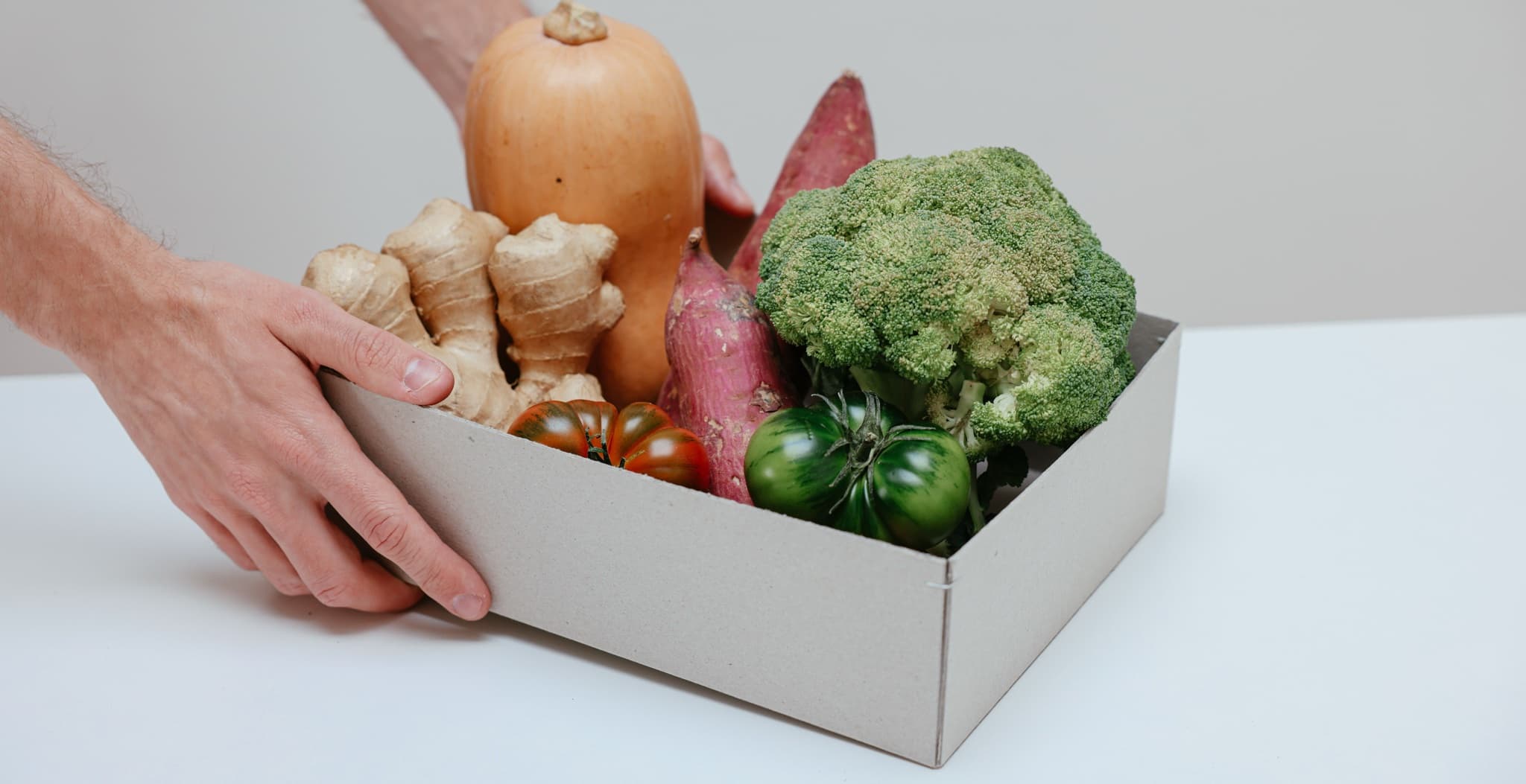Two hands placing a cardboard box packed with squash, ginger, sweet potatoes, broccoli, and tomatoes onto a white tabletop.