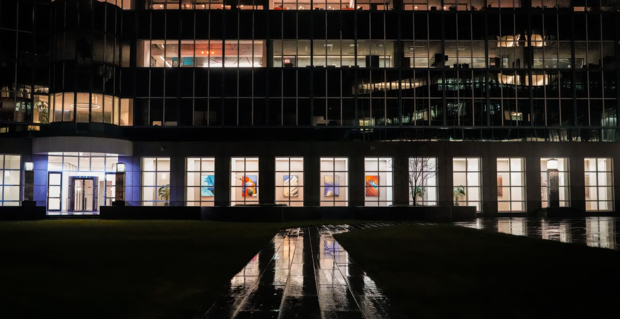 A glass-fronted office building lit up at night, with colorful abstract framed artwork visible through the ground-floor windows and a wet reflective walkway leading to the entrance.