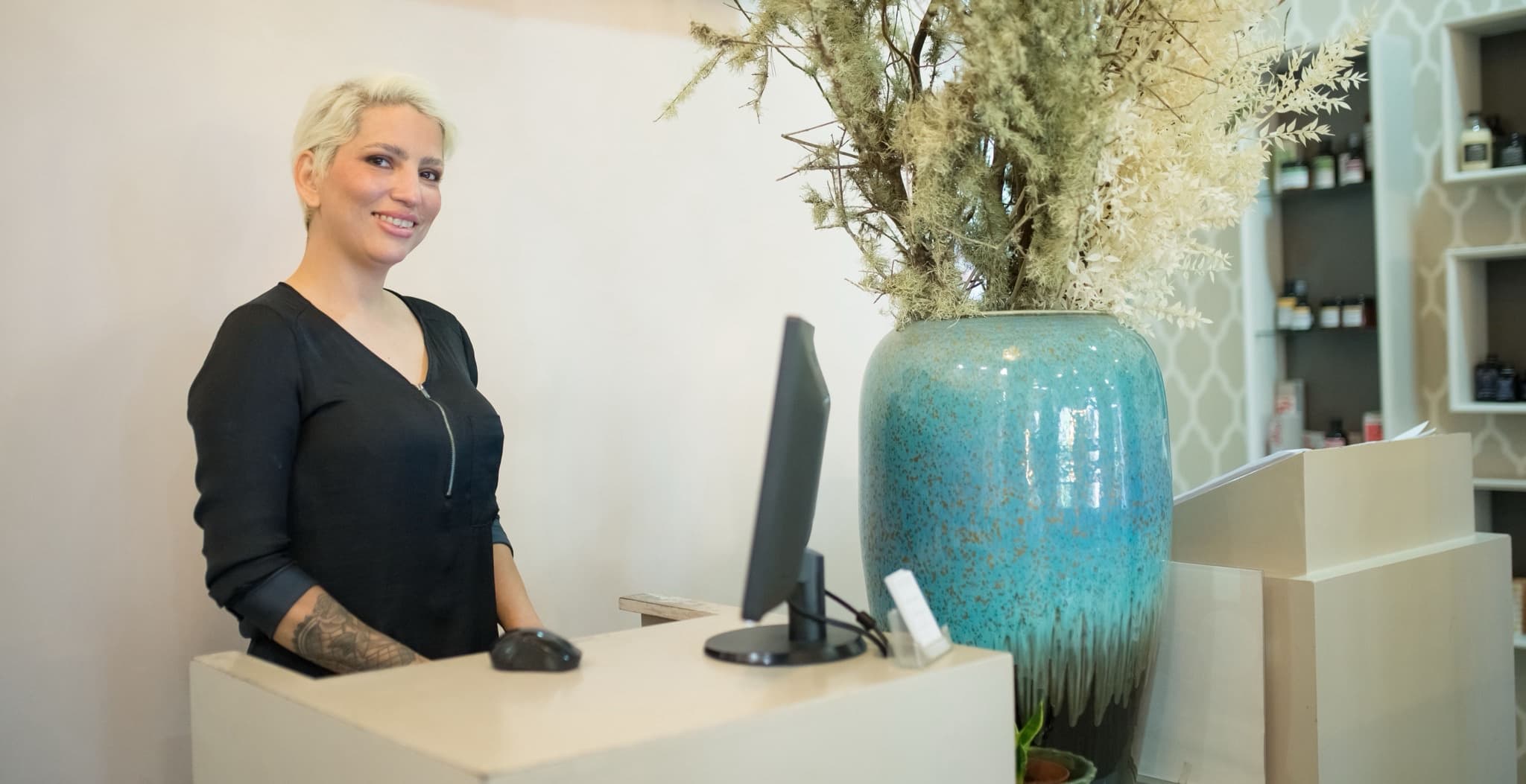 Smiling receptionist at a wellness studio front desk with a desktop computer and a large teal vase of dried botanicals