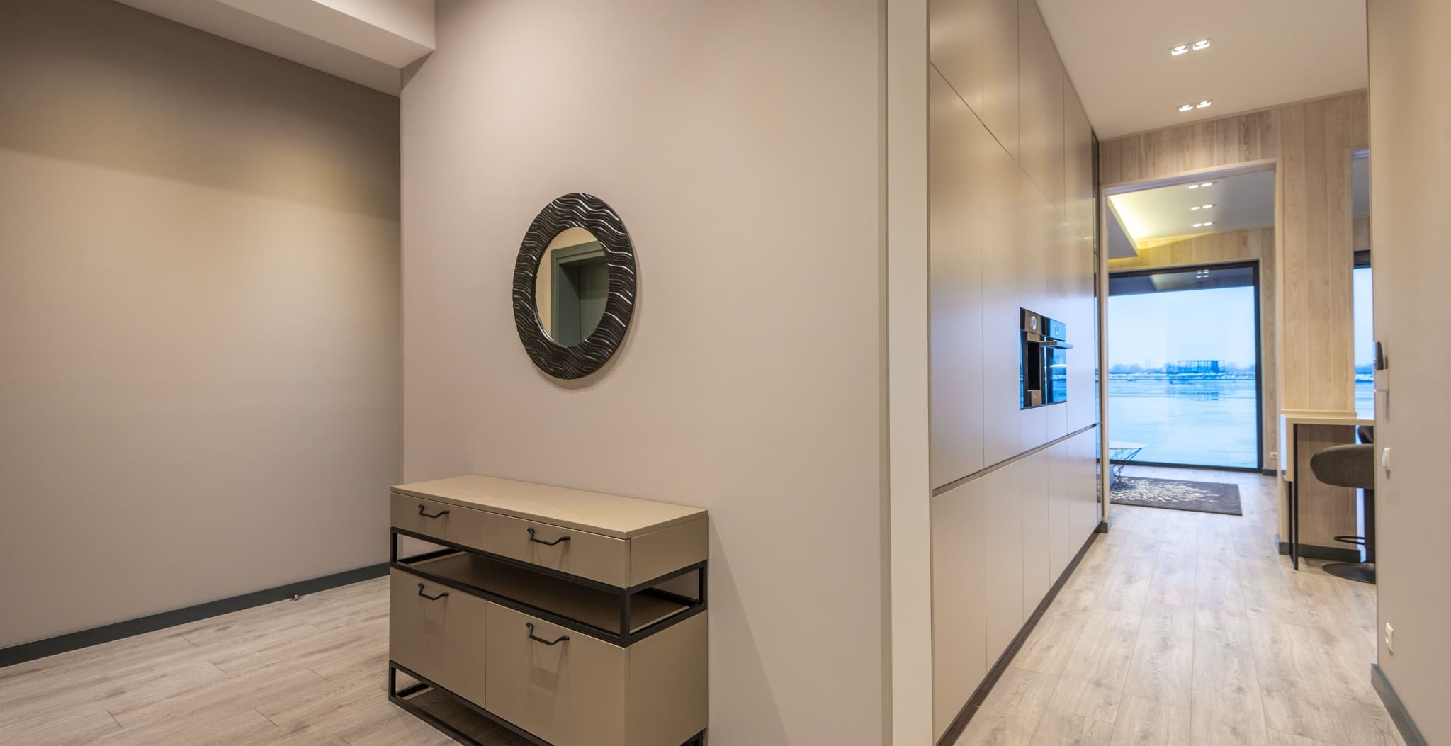 A minimalist hallway with a round wave-patterned mirror over a two-drawer console table, leading past built-in cabinetry to a glass door overlooking a frozen waterfront.