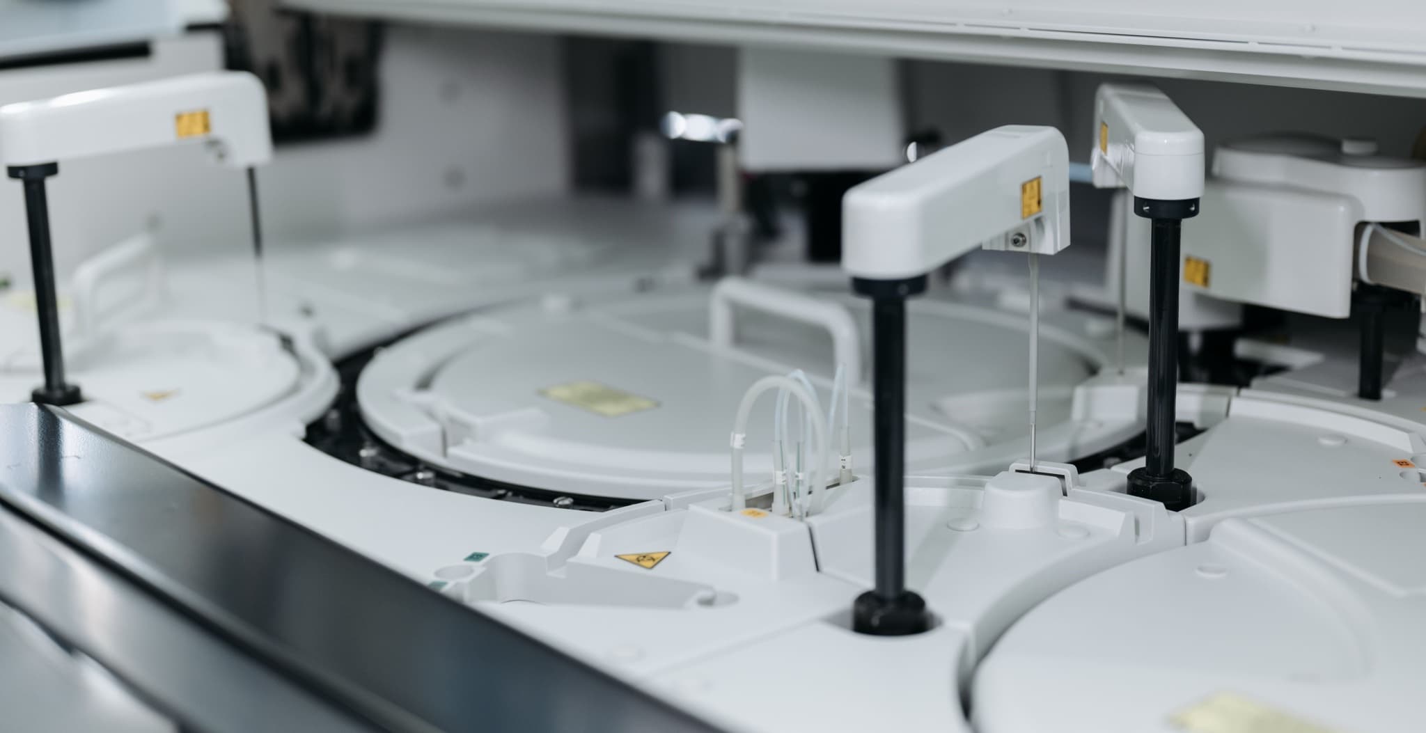 A close-up of white laboratory automation equipment with robotic dispensing arms, tubing, and yellow biohazard warning labels; no people are visible.
