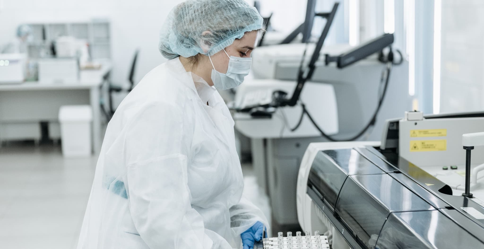 Scientist in a protective coverall, bouffant cap, mask and blue gloves carrying a rack of sample tubes toward a large benchtop laboratory analyzer instrument