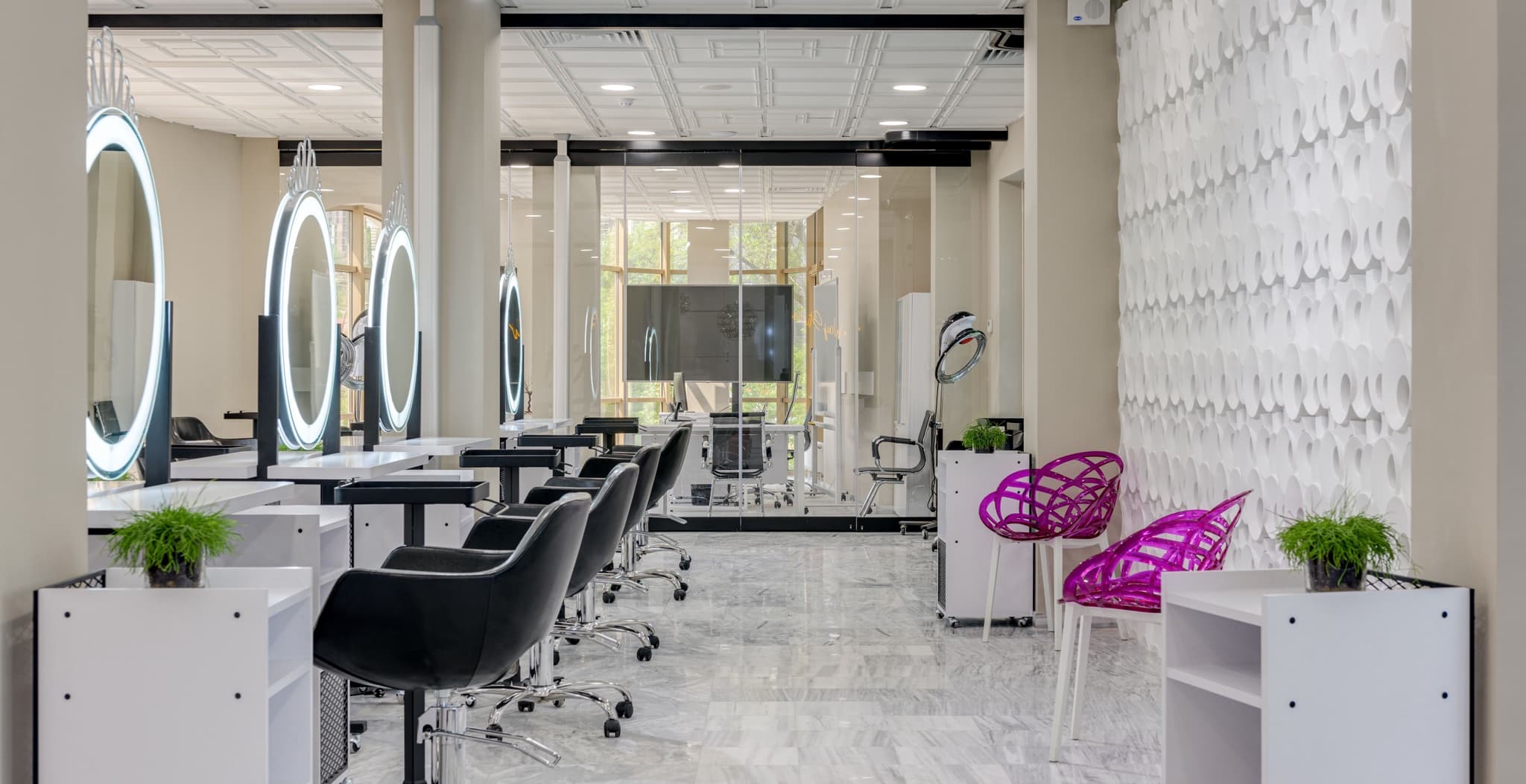 An empty upscale hair salon interior showing a row of black styling chairs beneath round lit mirrors, a marble floor, and pink accent chairs near a textured white wall.