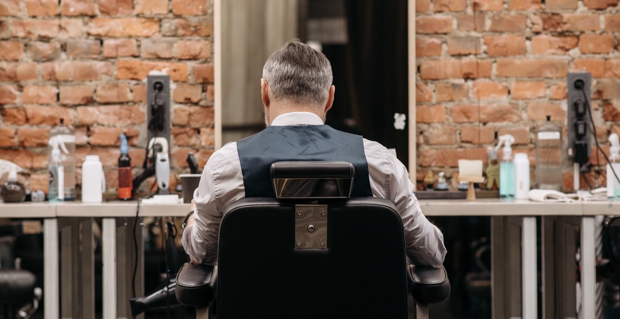 A rear view of a man with gray hair seated in a barber's chair facing an exposed brick wall lined with styling-product bottles and mirrors.