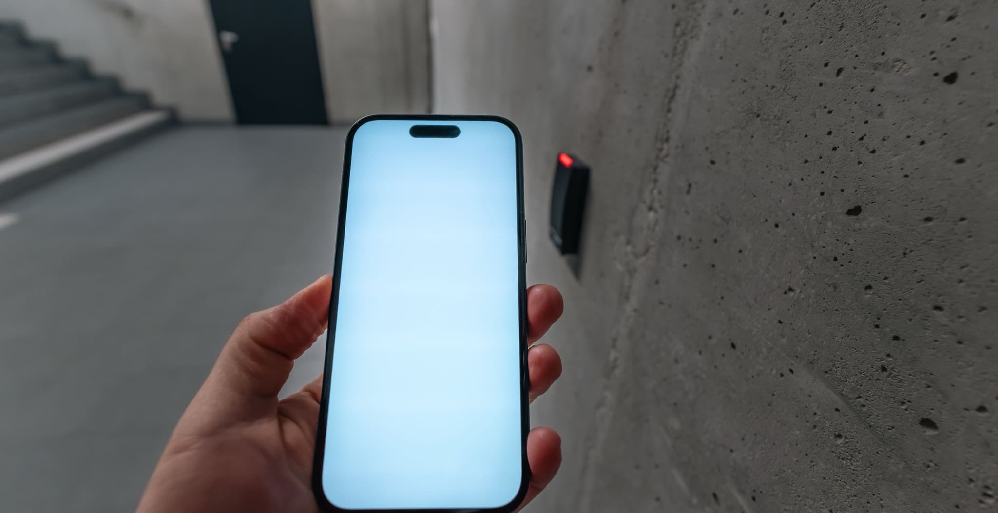 Close-up of a hand holding a smartphone with a glowing blank screen up to a wall-mounted door access reader (red indicator light) in a concrete stairwell corridor, no people's faces or branding visible