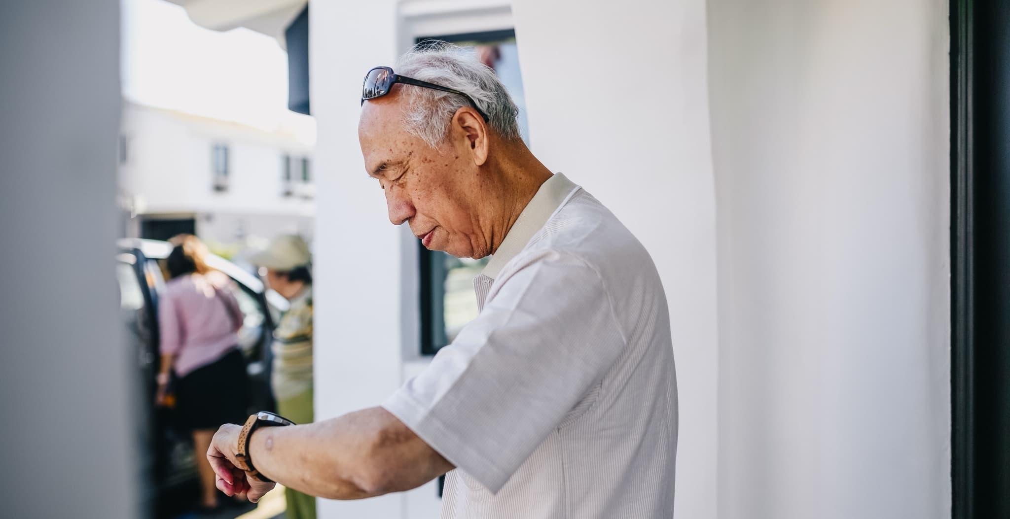 Senior man in a white shirt standing outdoors in bright daylight, glancing down to check the watch on his wrist