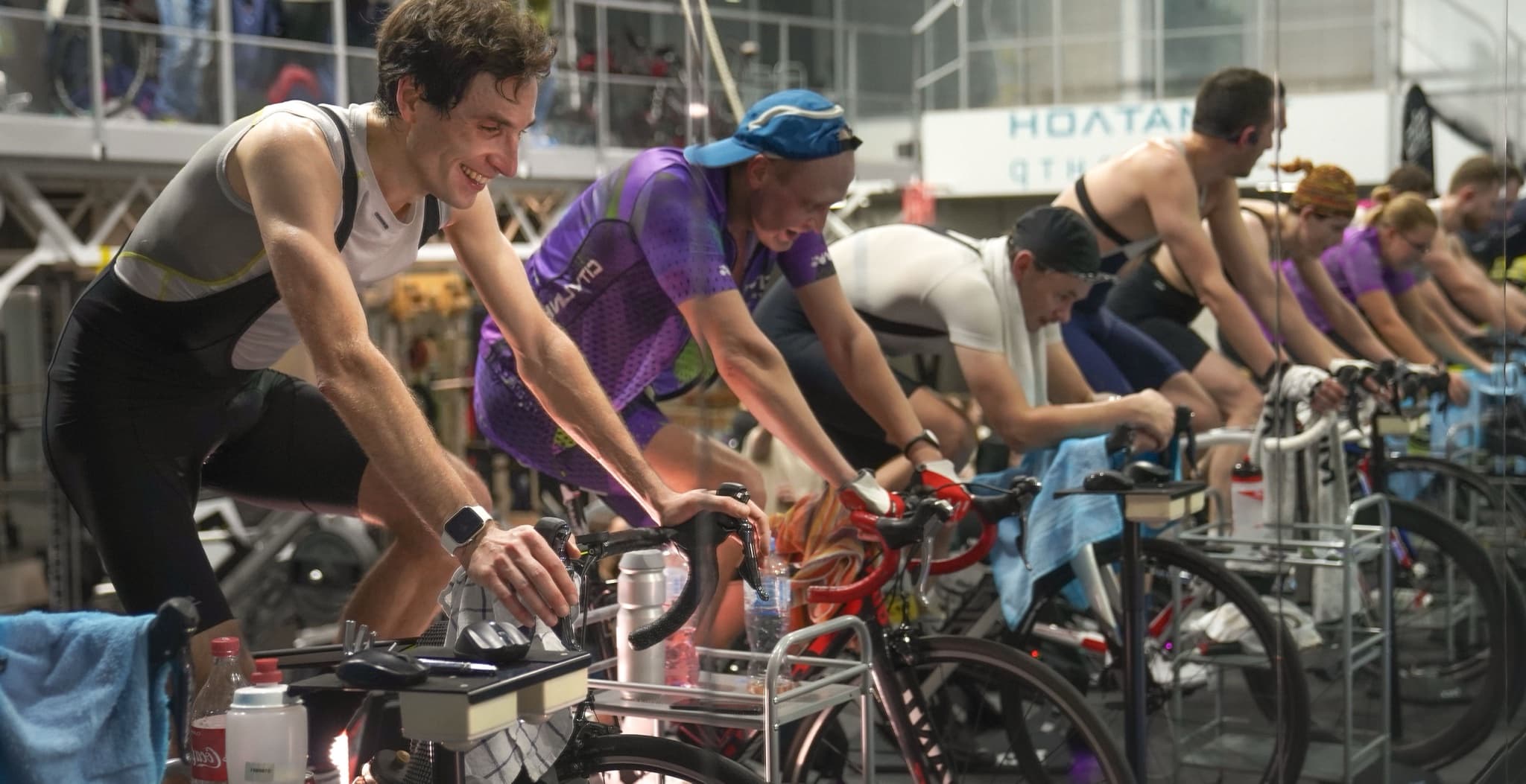 Row of members riding stationary bikes side by side in a dimly lit indoor cycling studio during a group training class