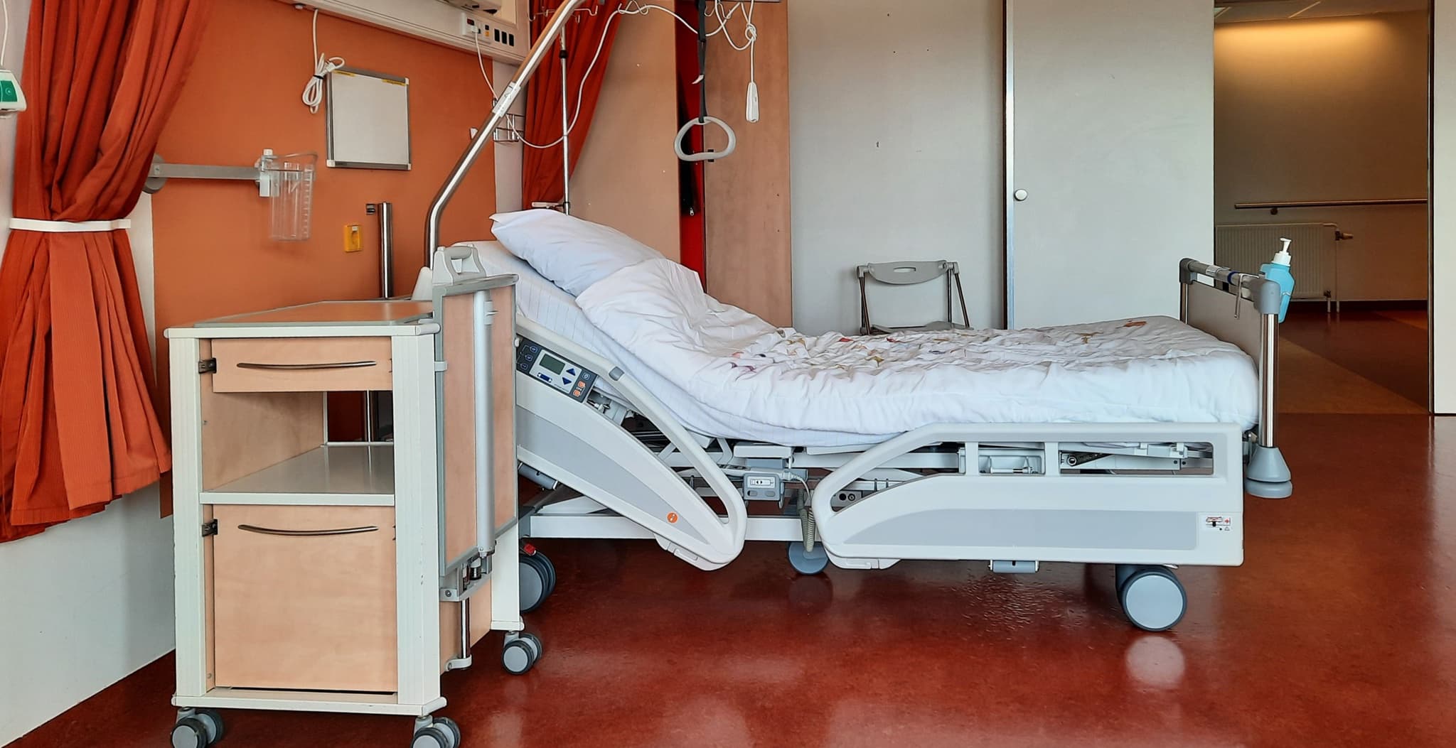 An empty adjustable hospital bed with white linens next to a wheeled bedside cabinet in a patient room with an orange accent wall and red privacy curtains.