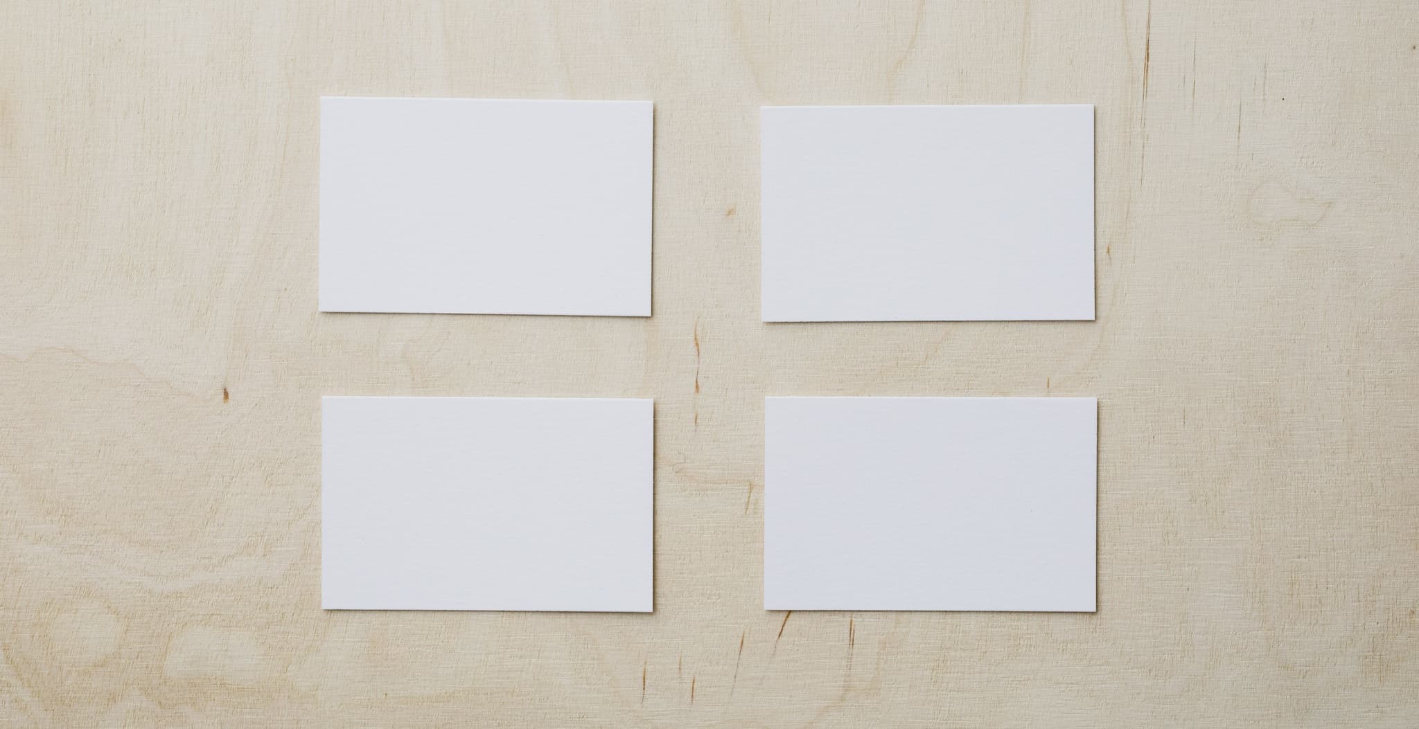 Four blank white business cards laid out in two rows on a light wood tabletop.