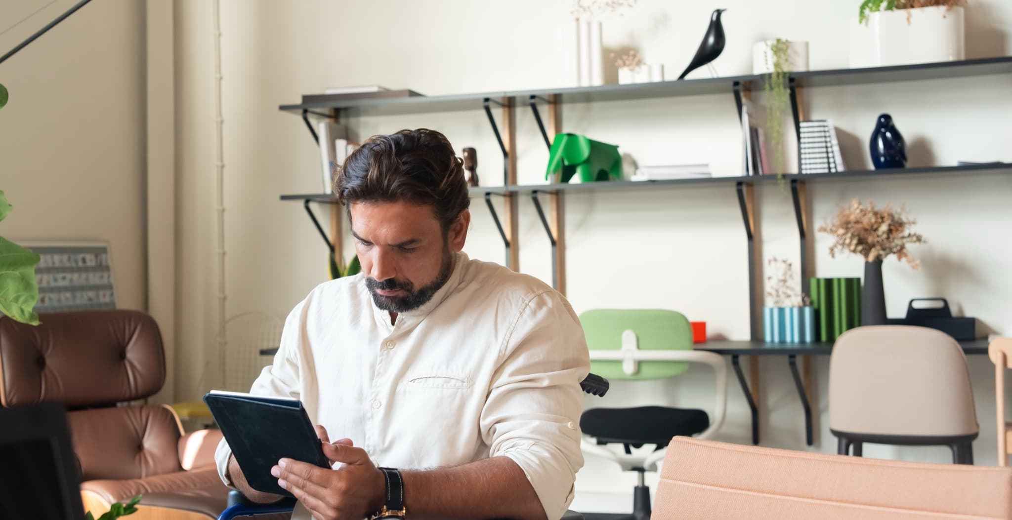 Man at a bright home-office desk browsing on a tablet held in both hands, with open shelving and chairs behind him